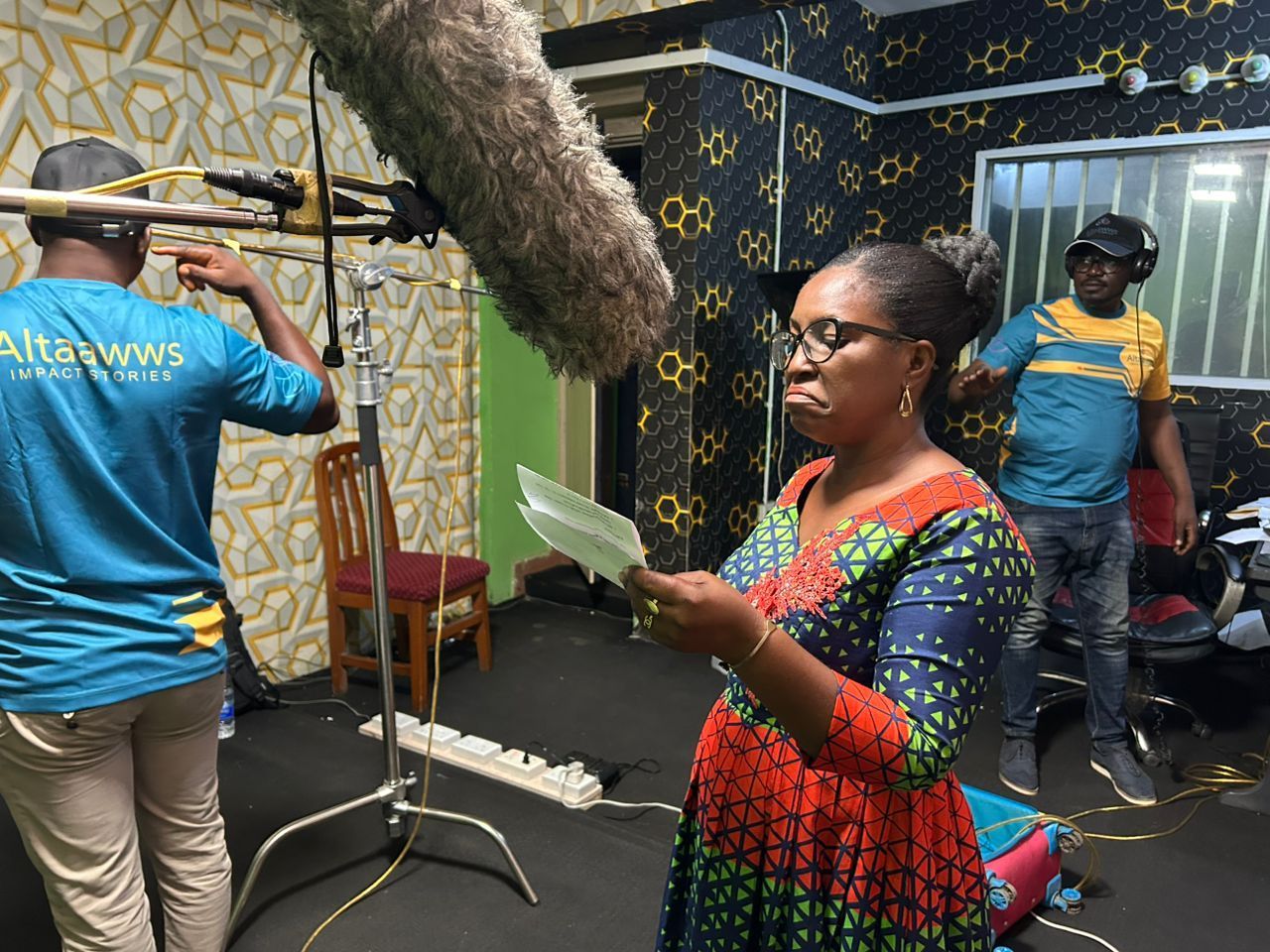 Woman in colorful dress reads script on film set, boom mic overhead. Crew adjusts equipment.