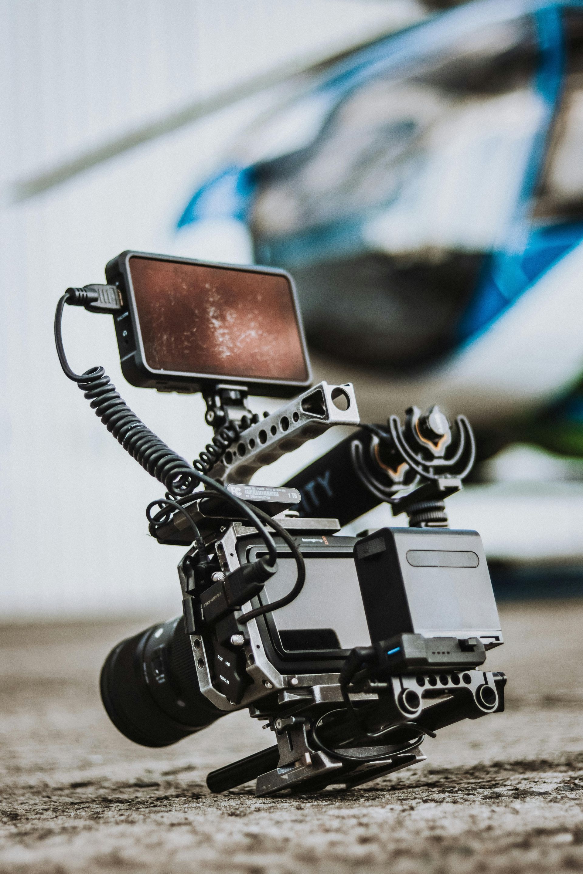 Professional video camera rig on a tripod, with a mounted monitor and microphone, outdoors on a sandy surface.