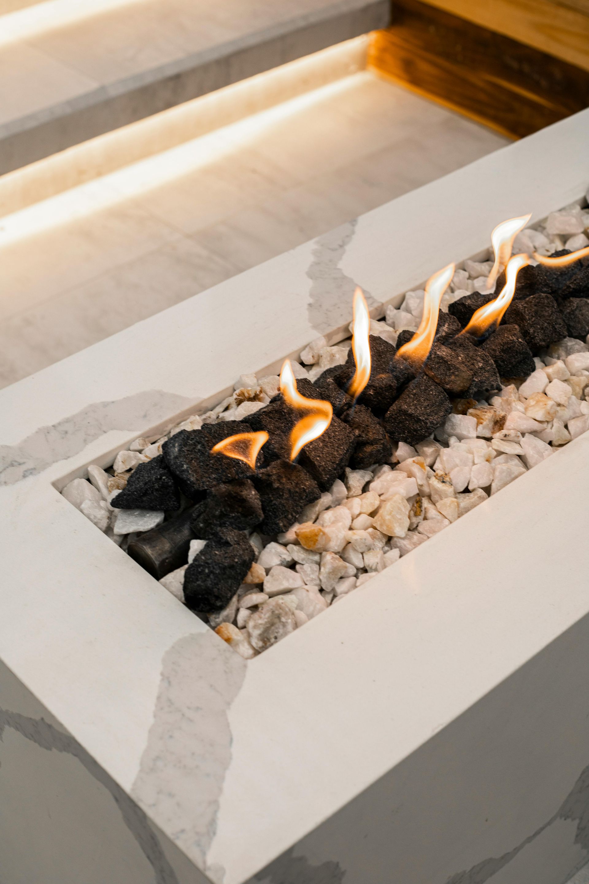 A rectangular, marble-patterned outdoor fire pit with flames flickering over black volcanic stones and white gravel.