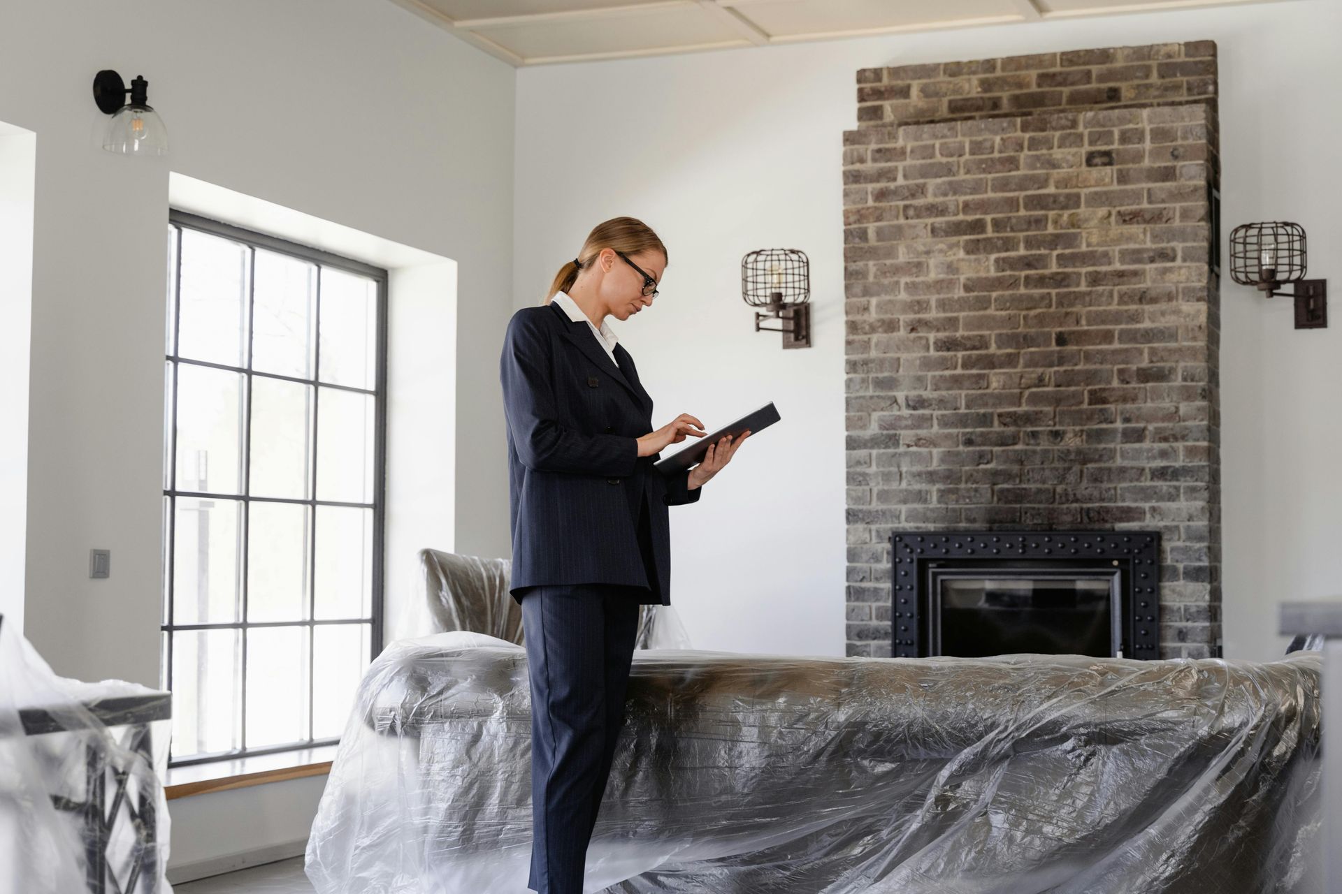 A person in dark clothes stands in a room under renovation, checking a digital tablet near a brick fireplace.
