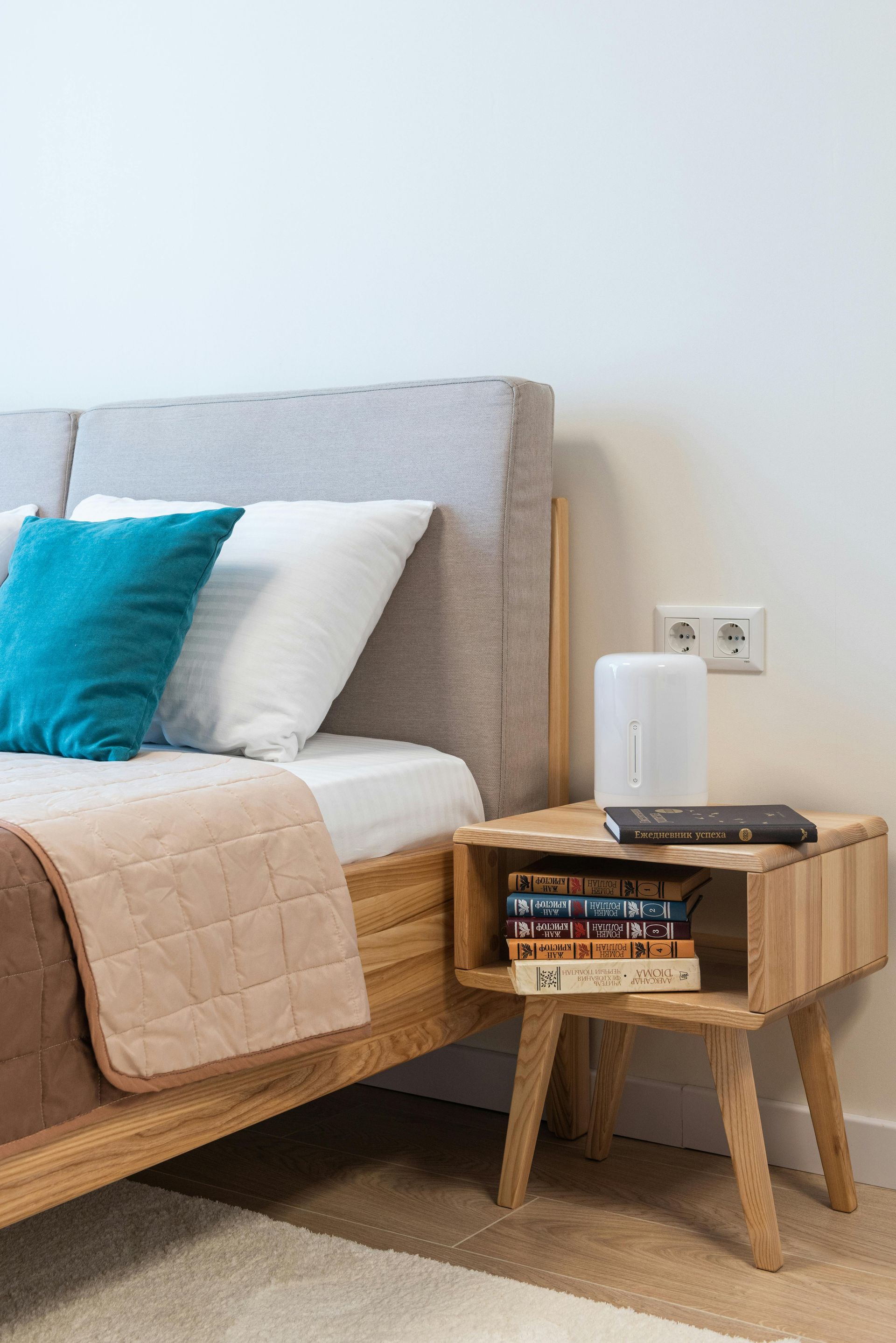 Bed with light wooden frame, pillows, blanket. Nightstand with books, lamp, outlet.