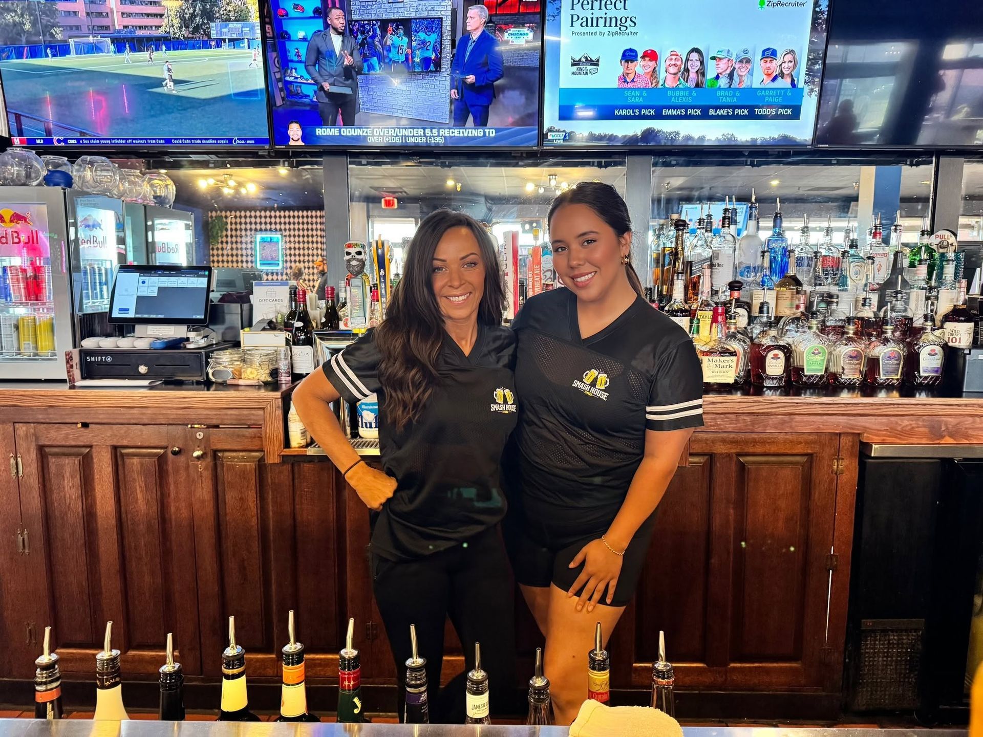 Two bartenders in black shirts standing behind a bar with bottles and TV screens in the background