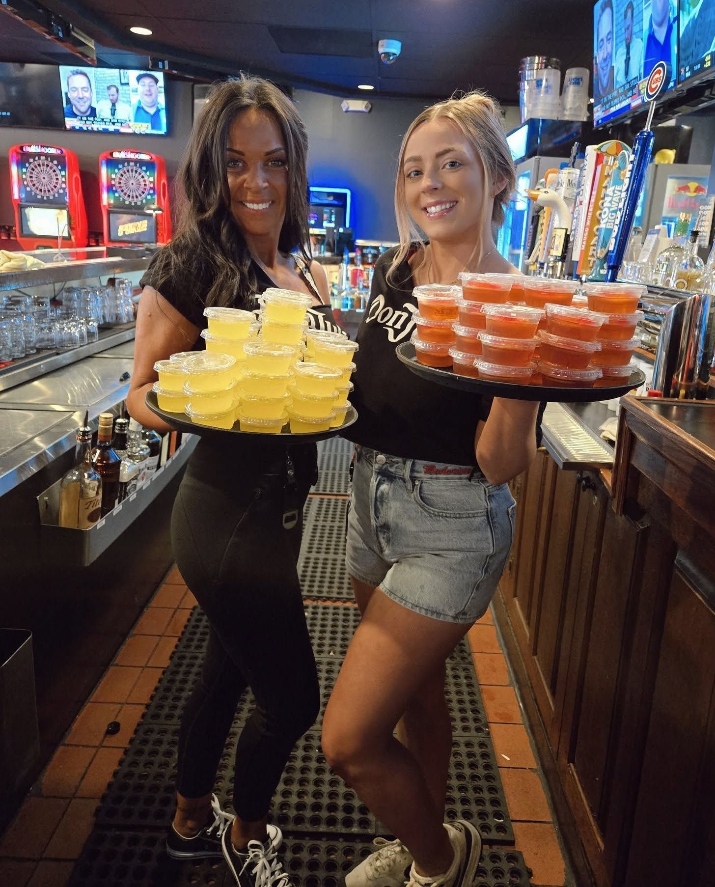 Two smiling bartenders holding trays of yellow and orange jello shots in a bar.
