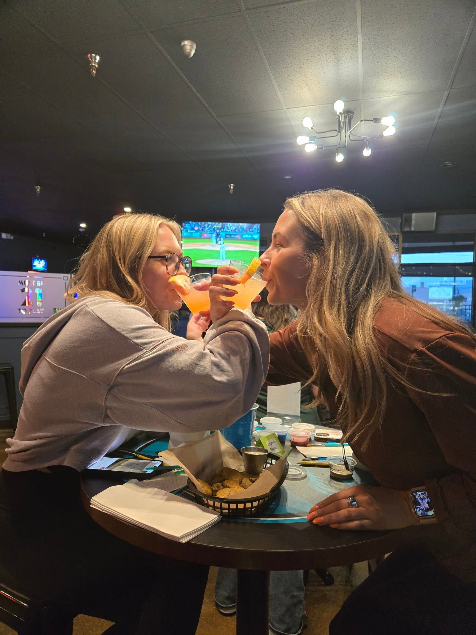 Two people leaning over a table, sharing a drink in a dimly lit bar with a TV in the background.