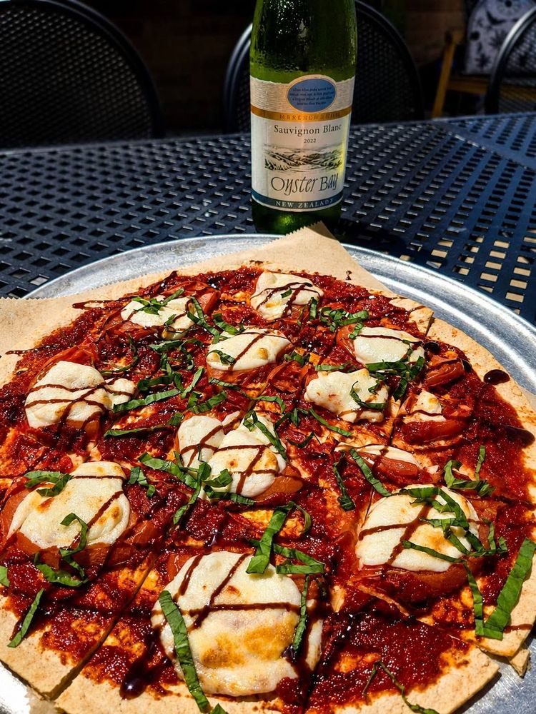 Pizza on a white plate with red sauce, cheese, herbs, and drizzled balsamic on a table, with a wine bottle behind it