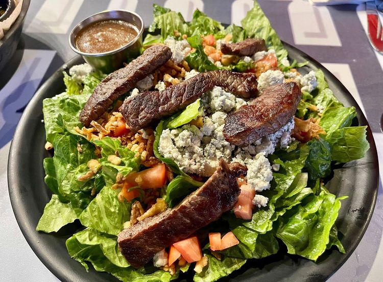 Grilled steak salad on a black plate with lettuce, tomatoes, and dressing on the side