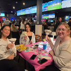 Three people toast at a pink table in a lively sports bar with multiple TV screens.