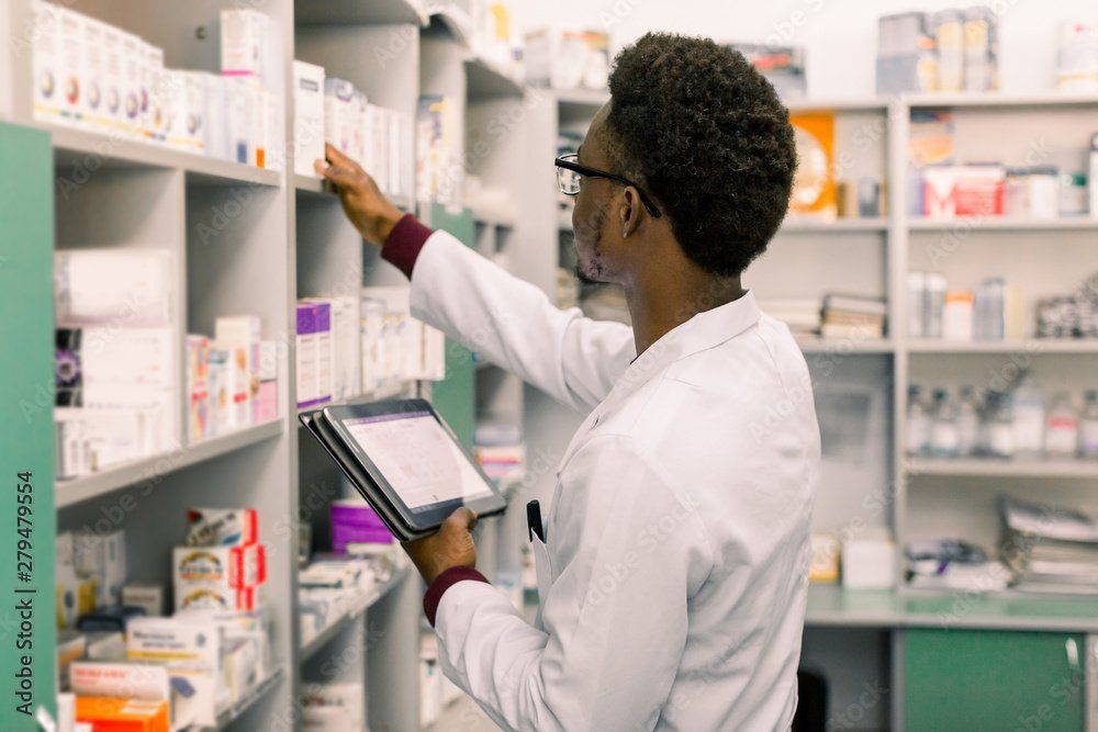 A pharmacist in a white coat checking medicine on a shelf while holding a digital tablet in a pharmacy.