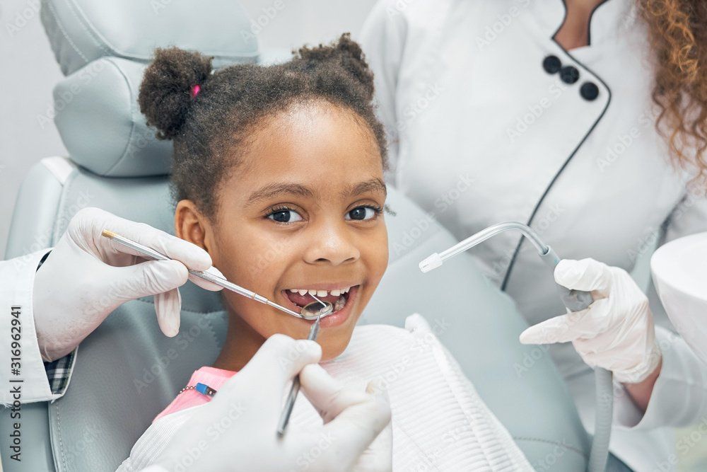 A dentist uses a mouth mirror and air-water syringe to examine a child sitting in a dental chair.