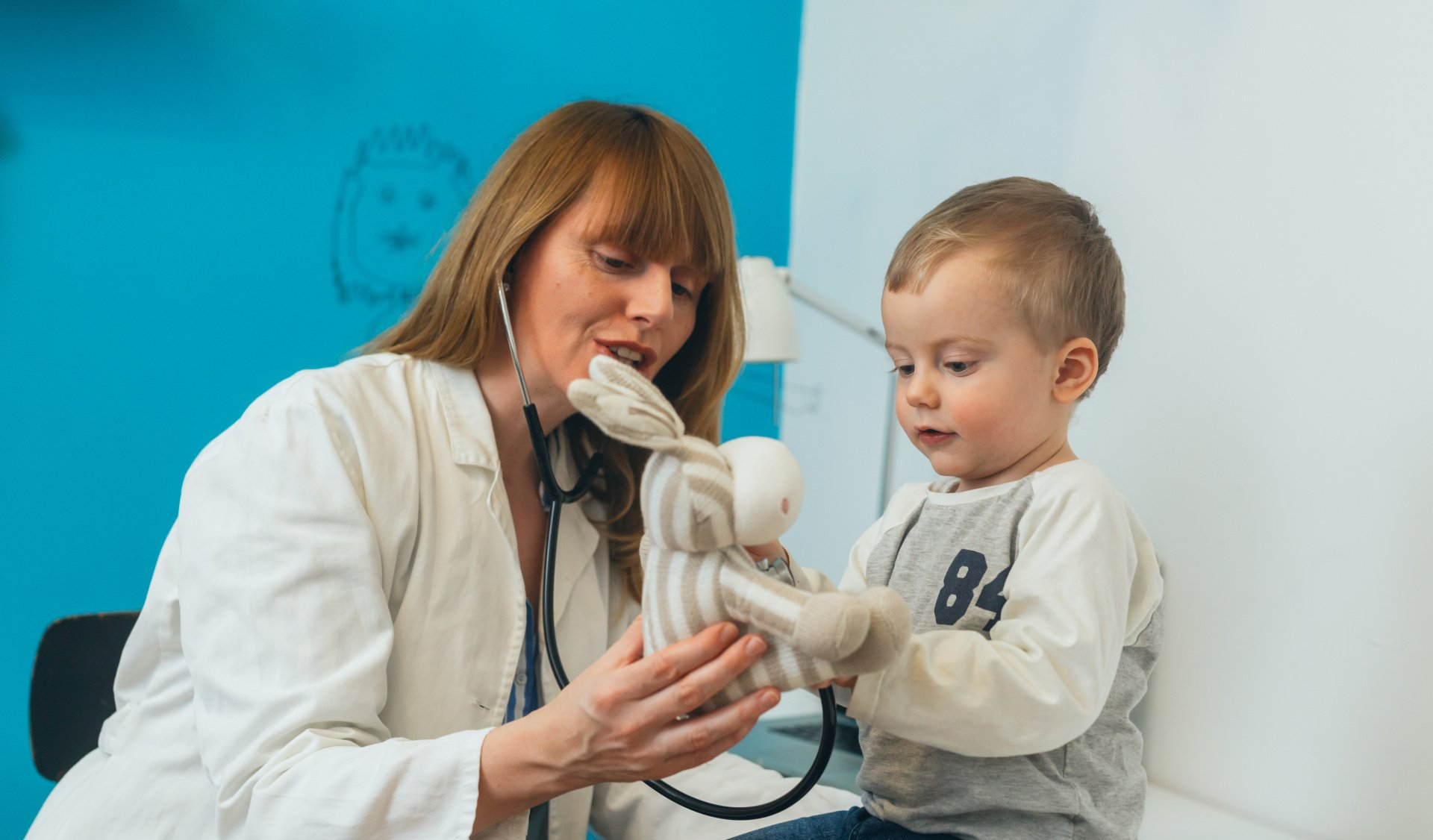 A doctor in a white coat uses a stethoscope to examine a plush rabbit toy held by a child in a doctor’s office.