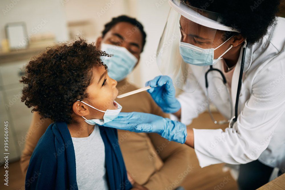 A medical professional in a lab coat and mask uses a wooden depressor to examine a patient's throat.
