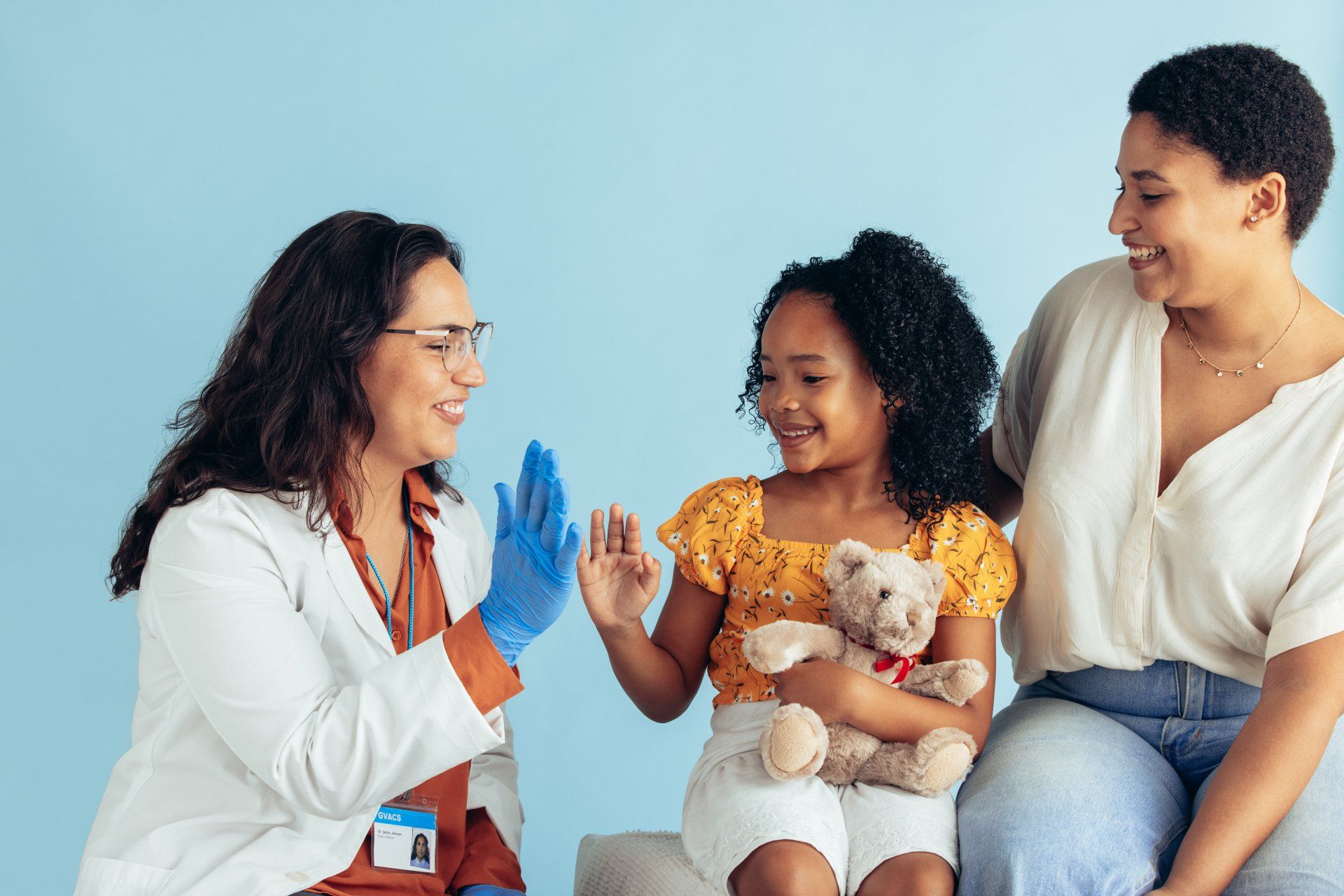 A doctor in a white coat and blue glove gives a high-five to a smiling child holding a teddy bear, next to a seated adult.