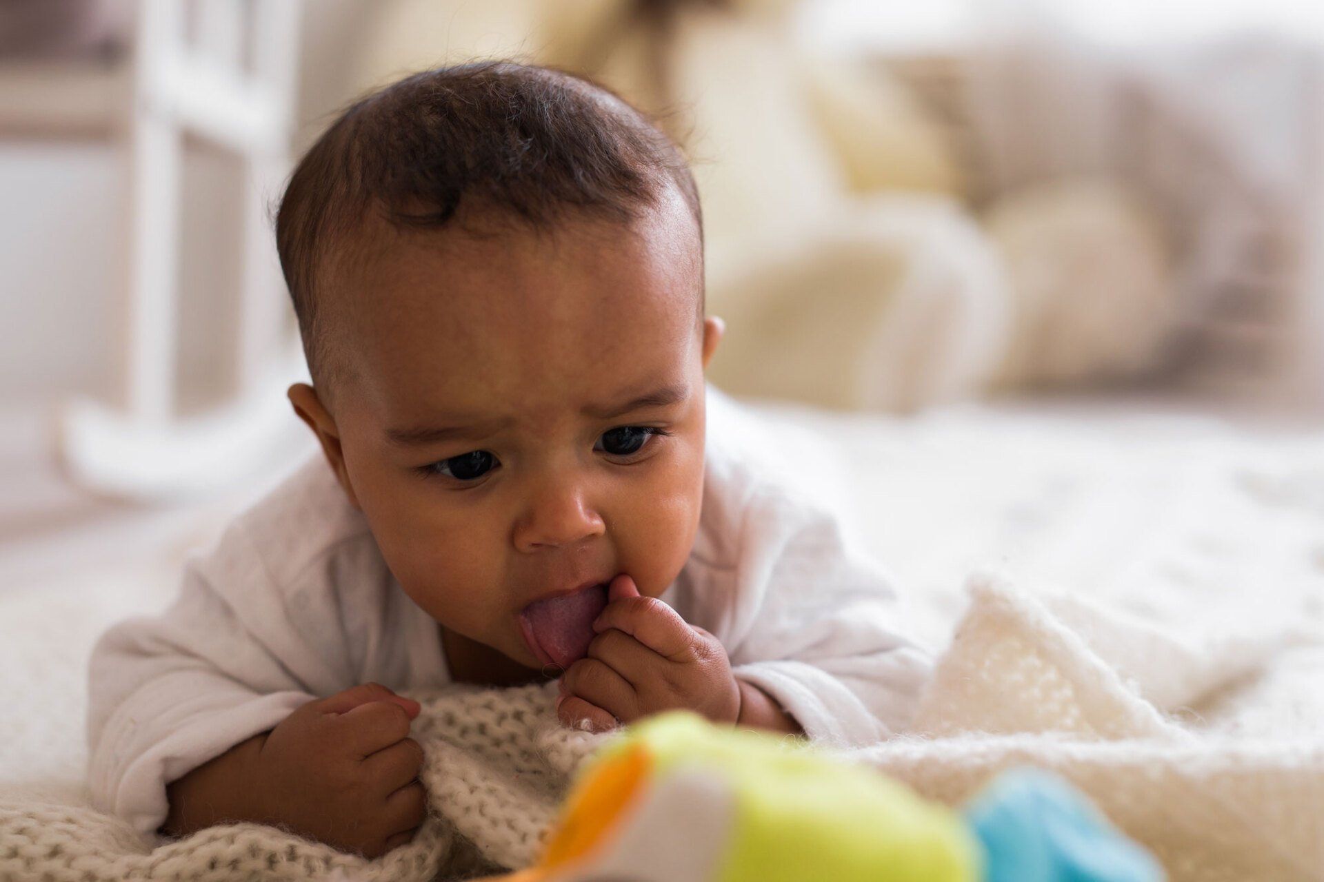 An infant on a white blanket explores a colorful toy, mouth open with a hand near its face in a soft-focus room.