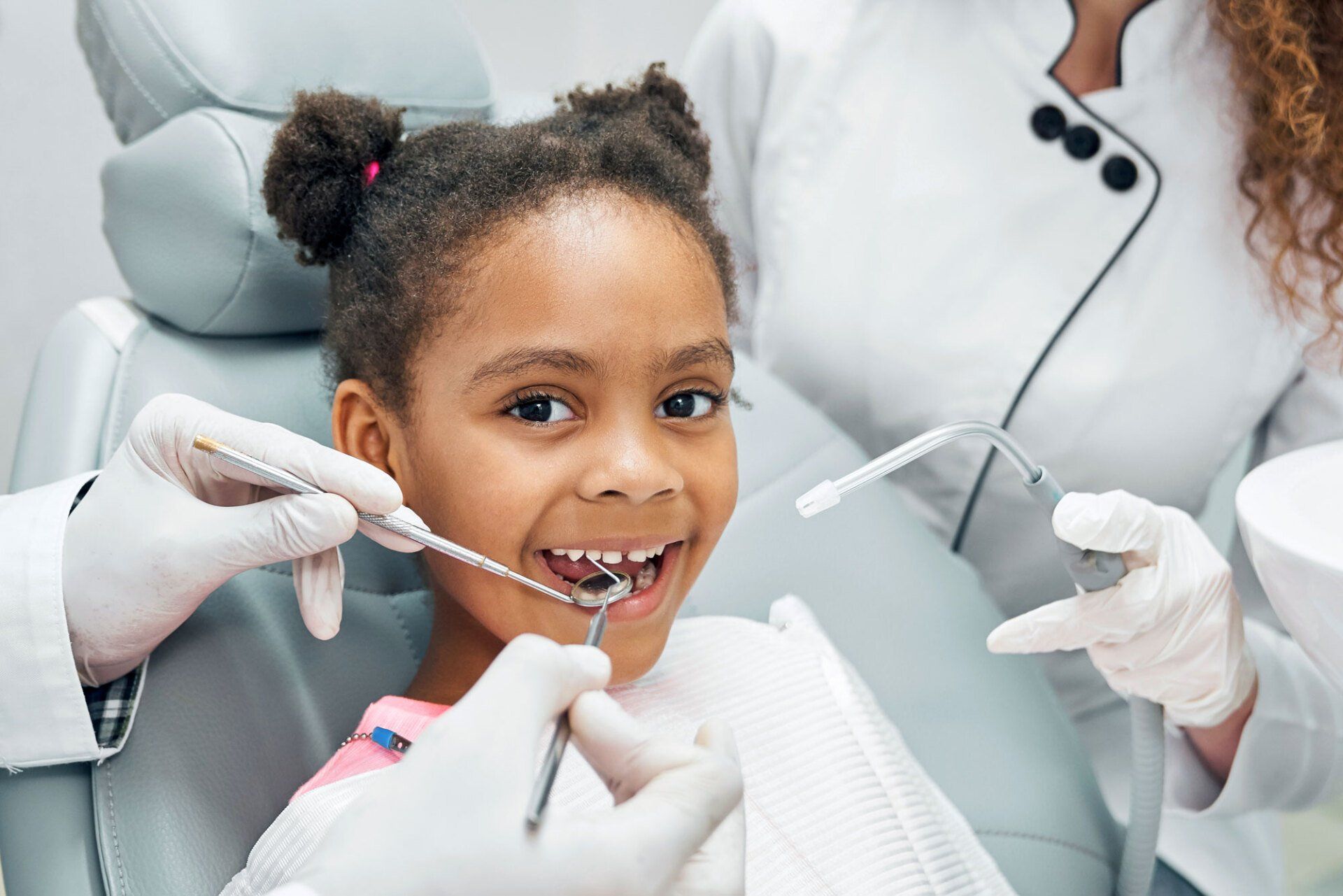 A child in a dental chair smiling while a dentist uses a dental mirror and suction tool during an exam.