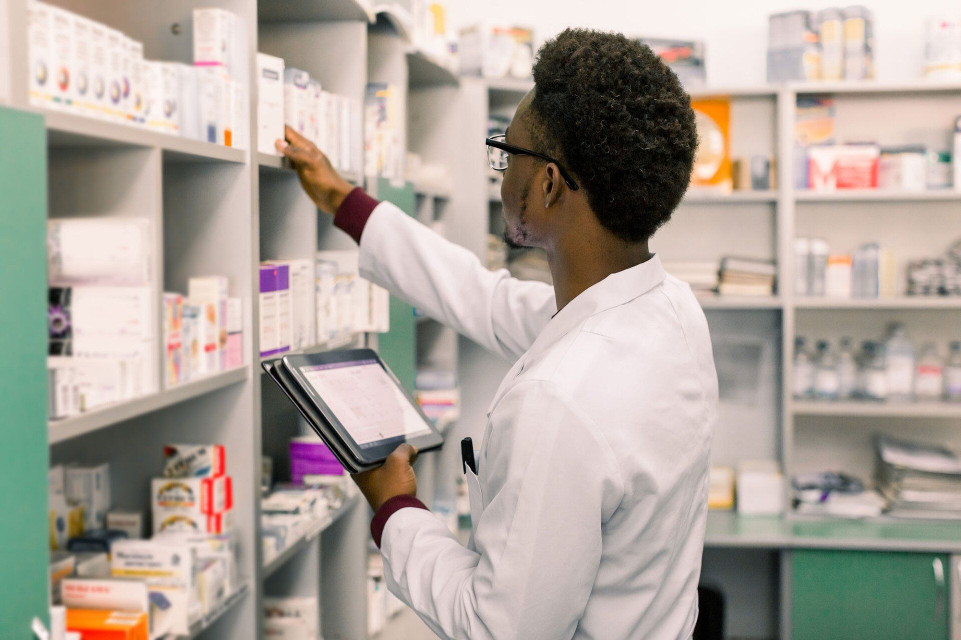 A pharmacist in a white coat reaches for medication on a shelf while holding a tablet in a pharmacy.