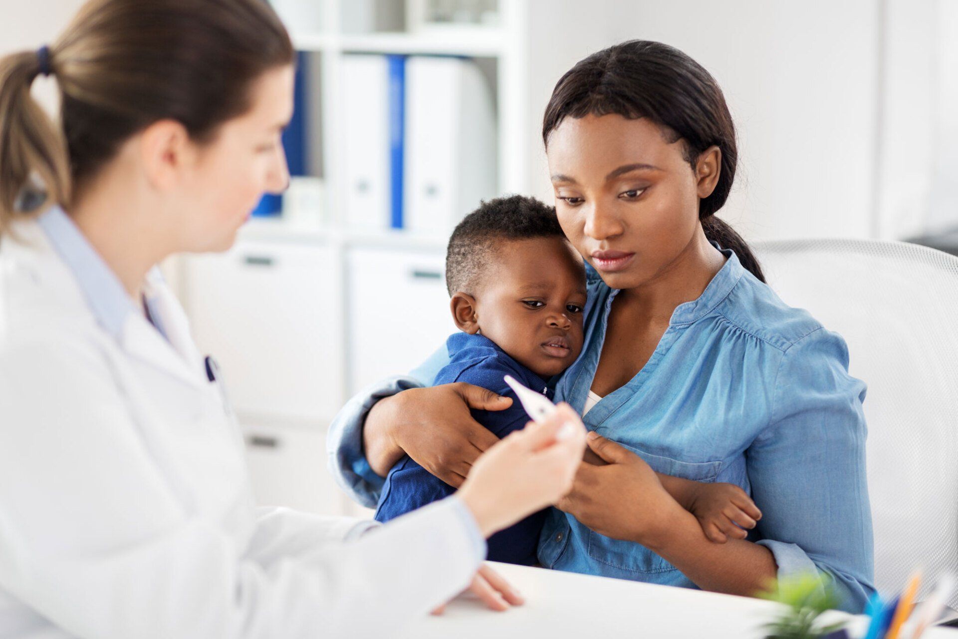 A doctor in a white coat shows a thermometer to a concerned parent holding a young child in a medical office.