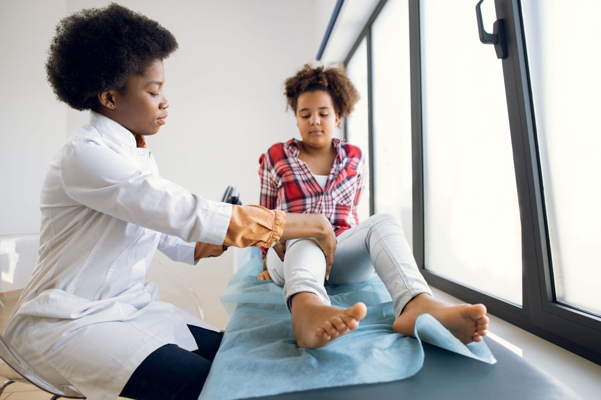 A doctor in a white coat examines a patient's knee in a clinical office setting.