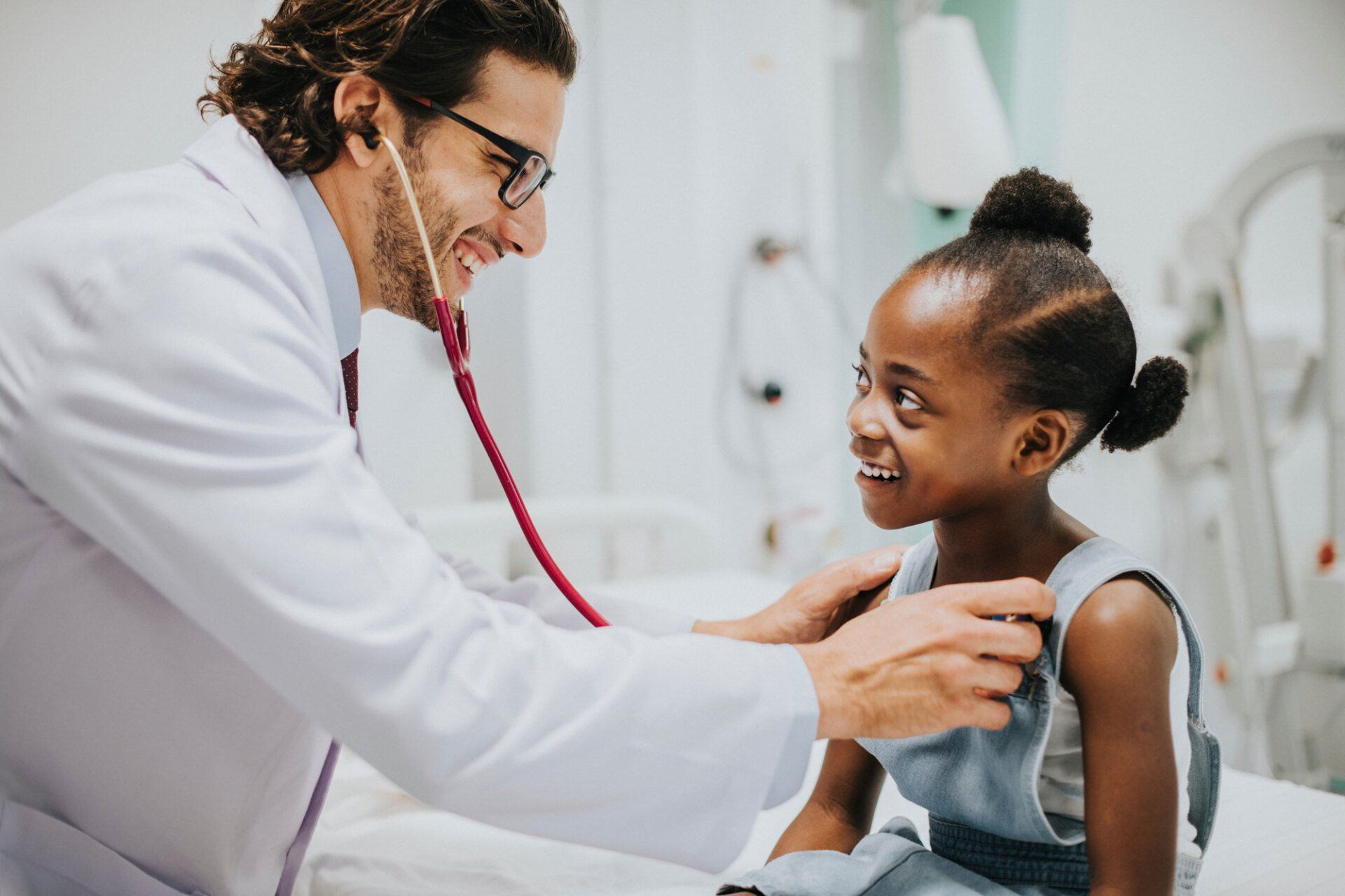 A smiling person in a white coat examines a patient with a stethoscope in a bright clinical setting.