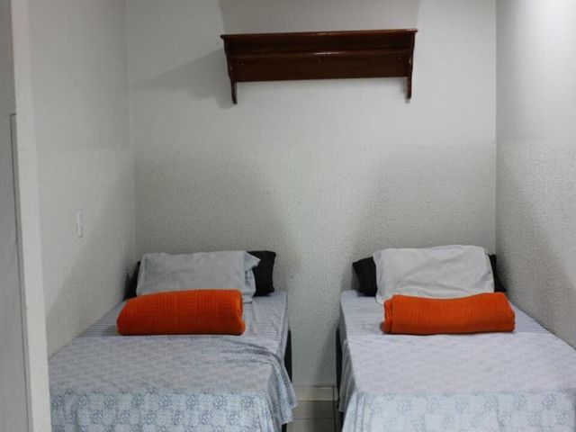 Duas camas de solteiro com lençóis brancos e travesseiros laranja em um pequeno quarto branco.