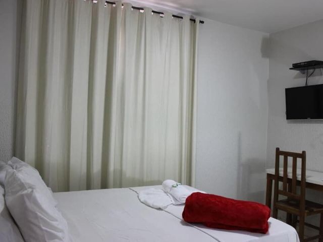 Quarto com cortinas brancas, cama desarrumada e TV fixada na parede ao lado de uma cadeira de madeira.