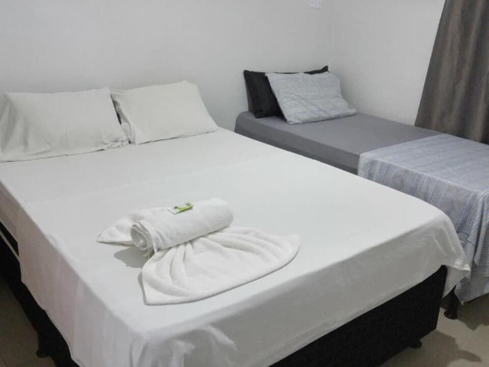 Duas camas arrumadas num quarto de hotel simples e branco, uma delas com toalhas dobradas por cima.