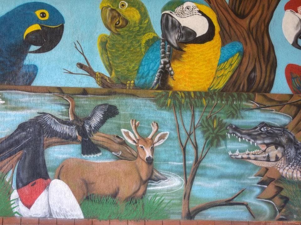 Mural colorido pintado com pássaros tropicais, um veado e um crocodilo entre galhos e água.