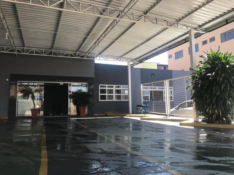 Entrada externa coberta com pavimento molhado, prédio escuro, plantas em vasos e corrimão de metal em forma de rampa.