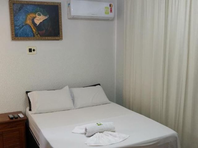 Cama arrumada num quarto simples com lençóis brancos, cortinas, mesa de cabeceira e obras de arte na parede.