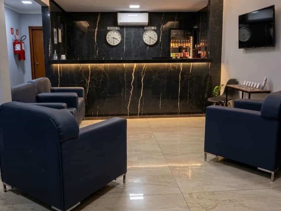 Hall de entrada moderno com cadeiras azul-marinho, balcão de recepção preto, relógios de parede e TV fixada na parede.