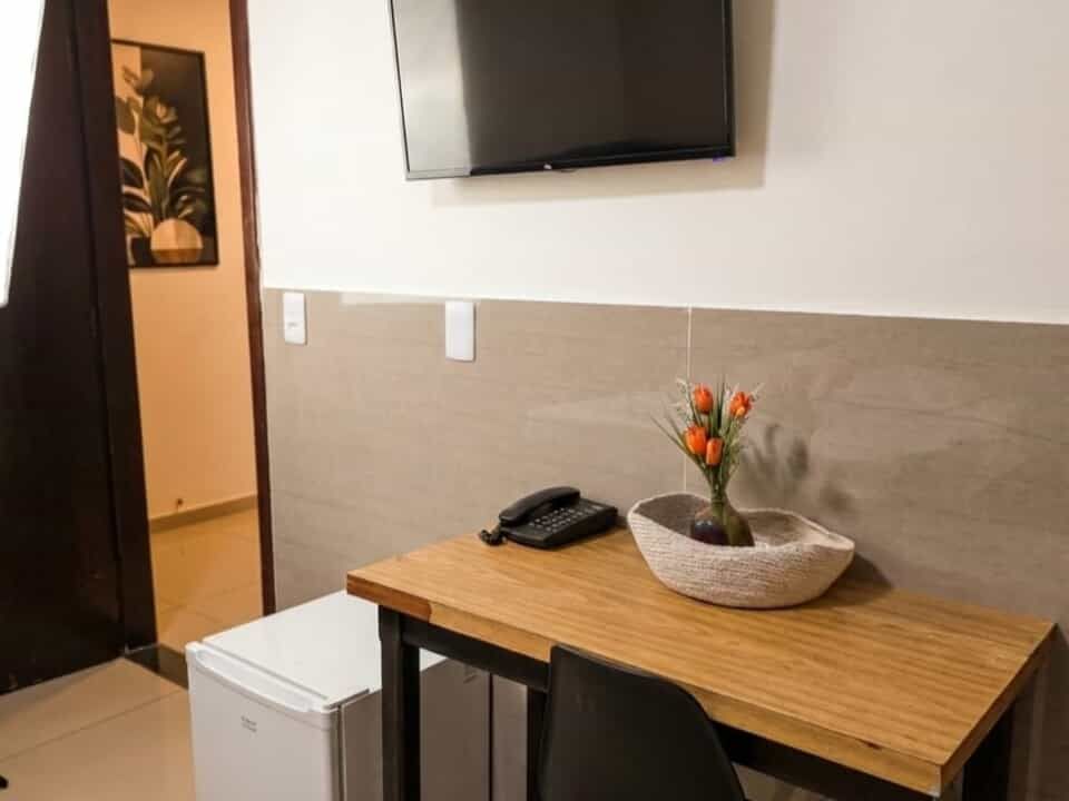 Quarto pequeno com TV de parede, escrivaninha de madeira, frigobar e vaso de flores laranjas.