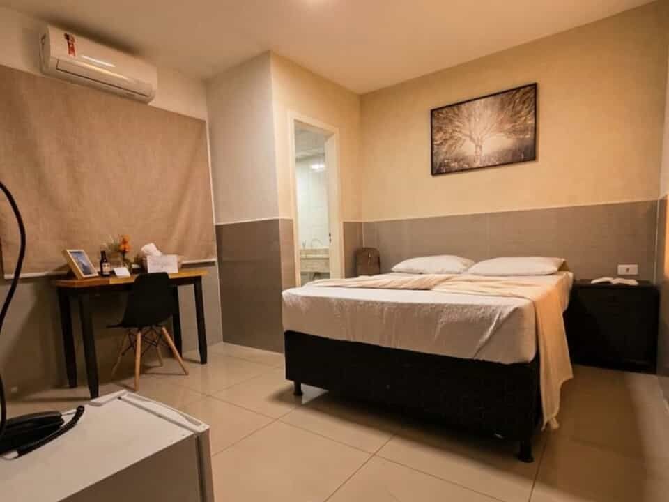 Quarto de hotel pequeno com cama de casal, escrivaninha, ar-condicionado e paredes bege.
