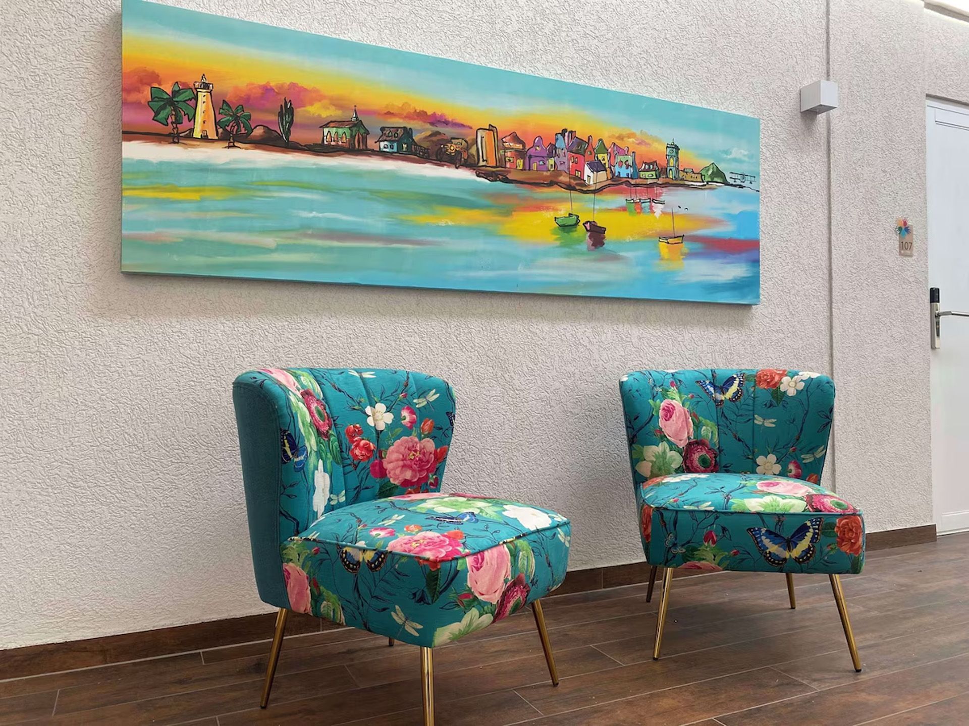 Dos sillas verde azulado con motivos florales debajo de una pintura de paisaje sobre una pared blanca con textura.
