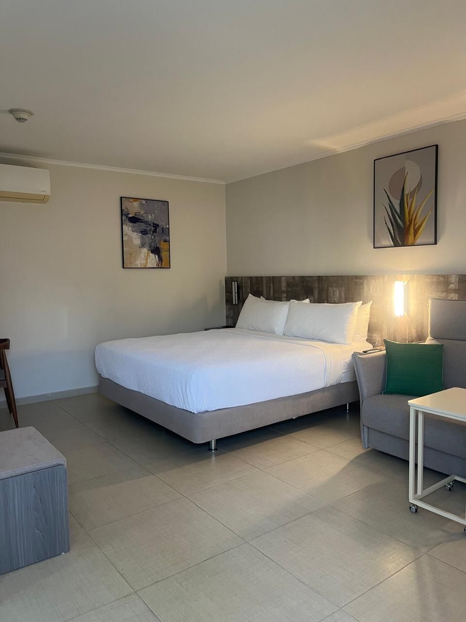 Dormitorio con cama, sofá, cuadros y una mesita. Tonos neutros, bien iluminado.