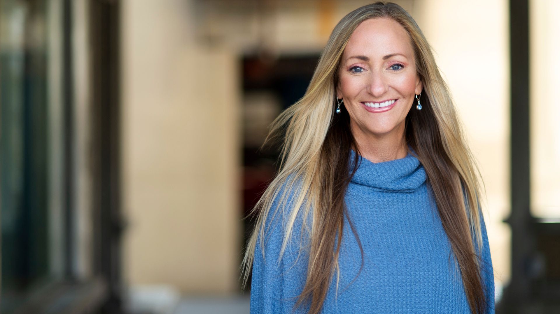 Smiling woman in a blue sweater standing in a blurred indoor hallway