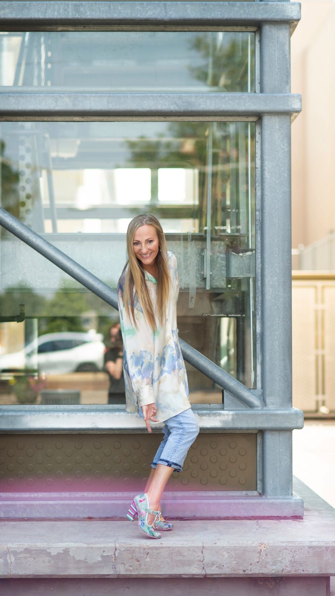 Woman standing on a step outside a glass building, wearing a light cardigan and jeans.