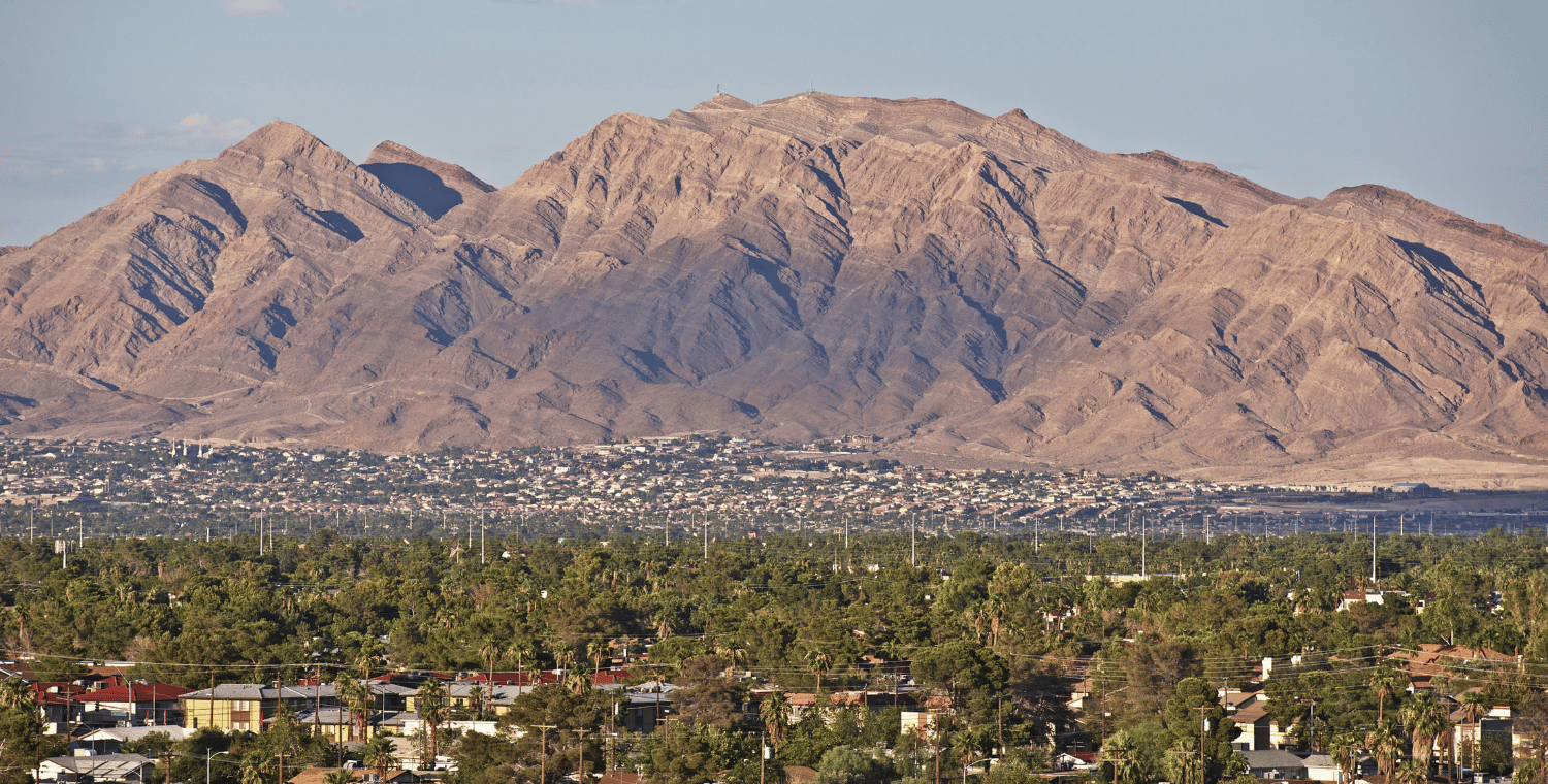 Brown mountains rise over a city with green trees in the foreground.