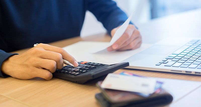 Person using calculator, with paperwork and credit cards on desk, near laptop.