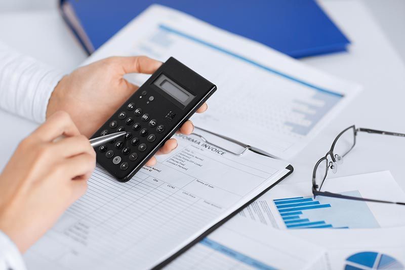 Hands using a calculator and pen to review financial documents with charts and a blue binder on a desk.