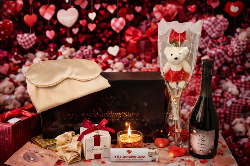 Valentijnsdag cadeauset: berenboeket, champagne, chocolaatjes, slaapmasker, kaars, tegen een rode hartvormige achtergrond.