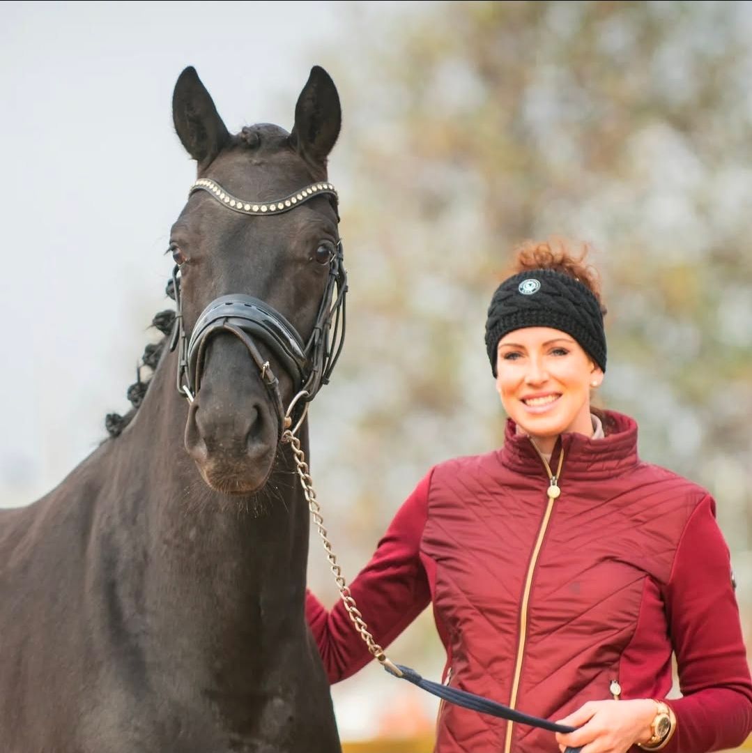 Een vrouw in een bordeauxrood vest glimlacht en houdt buiten een zwart paard aan de teugel.