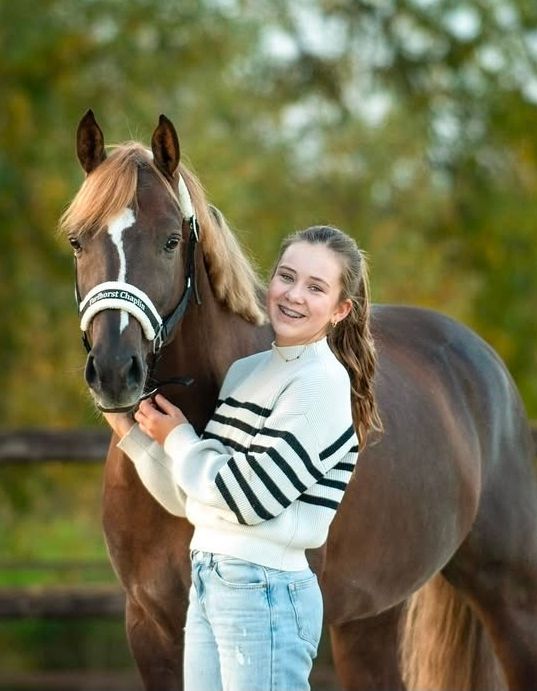 Een persoon met lang haar glimlacht terwijl hij in een veld naast een donkerbruin paard met een witte streep op zijn gezicht staat.