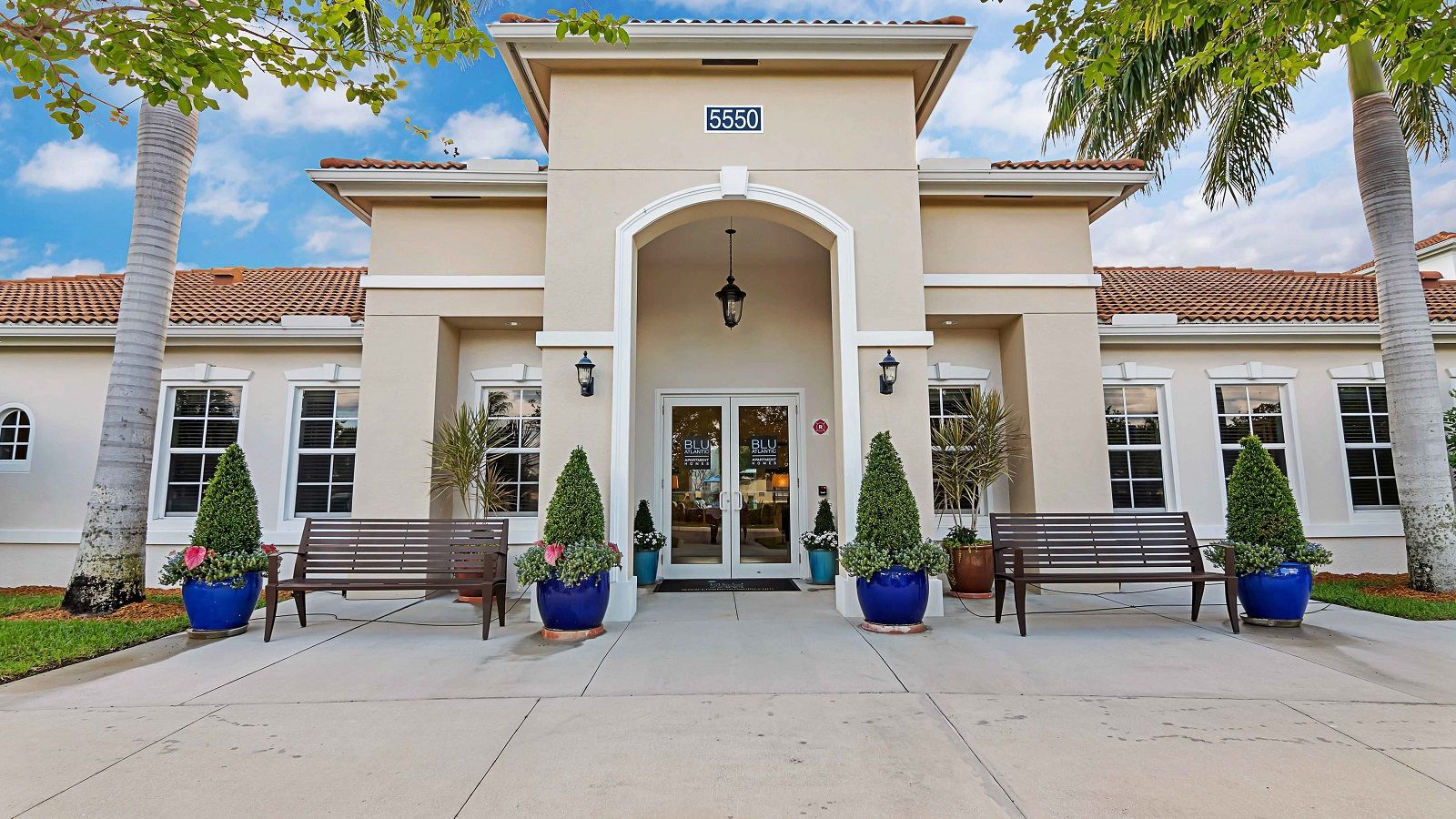 Modern white stucco building entrance with arched doorway, benches, potted plants, and palm trees