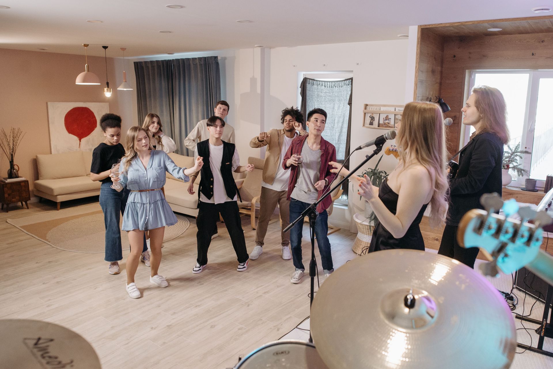 Musicians rehearsing in a bright living room, with guitars, mic stands, and a drum kit in the foreground