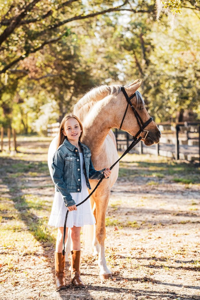 Young rider standing beside a saddled cream horse on a sunlit wooded path