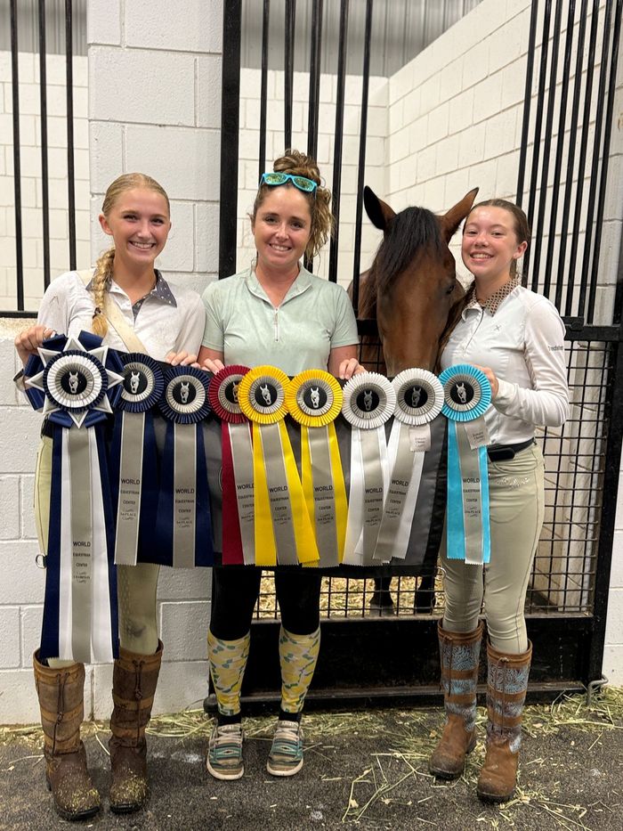 Three women hold horse show ribbons beside a dark horse in a barn aisle.