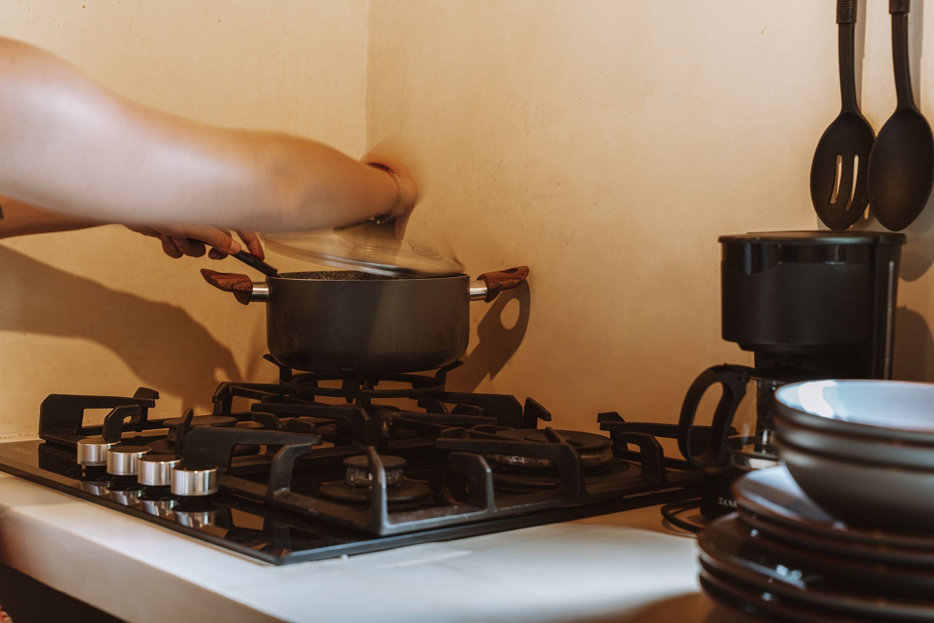 Hand stirring a black pot on a gas stovetop with stacked dishes nearby
