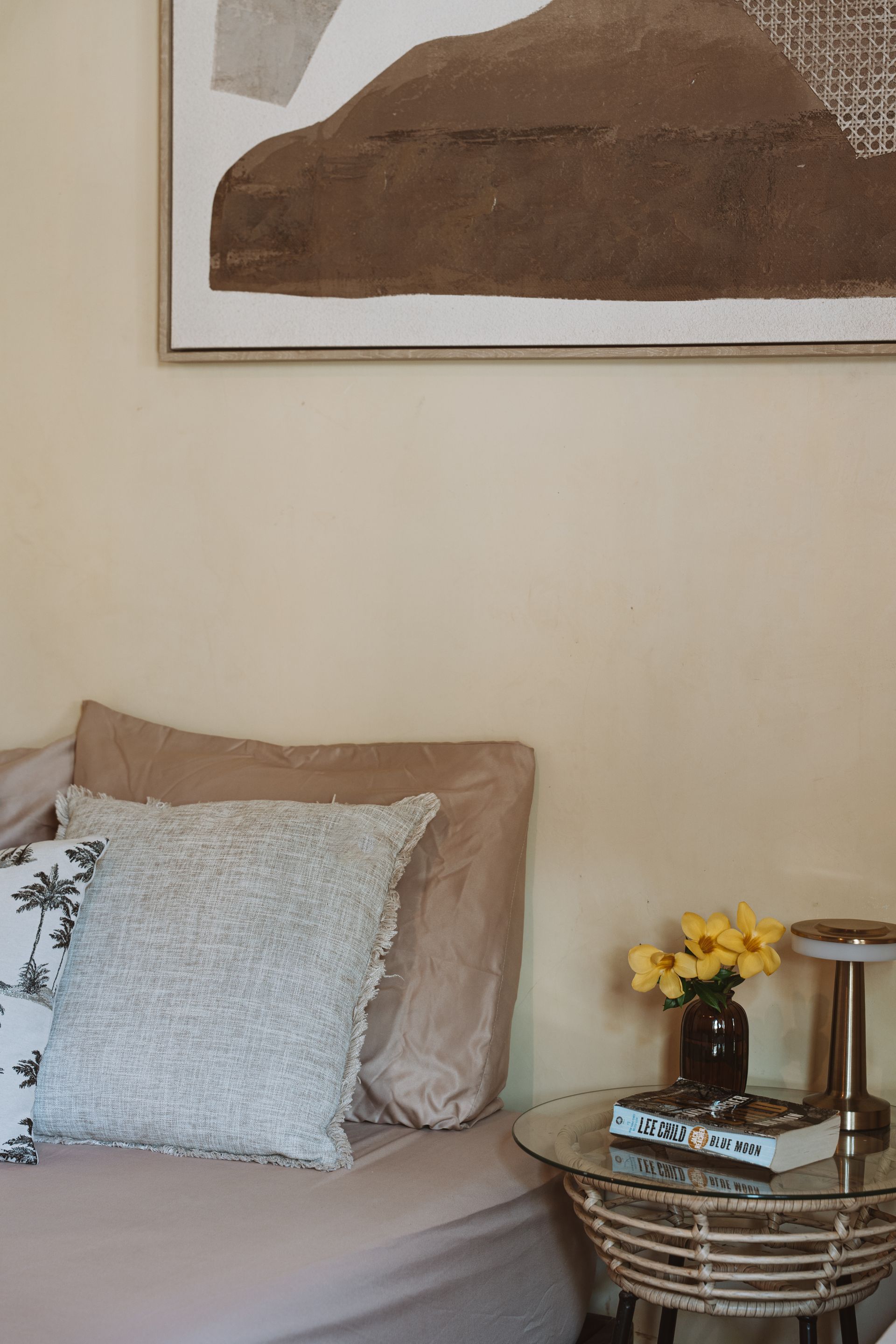 Cozy bedroom corner with beige bed, patterned pillow, woven side table, and framed wall art.