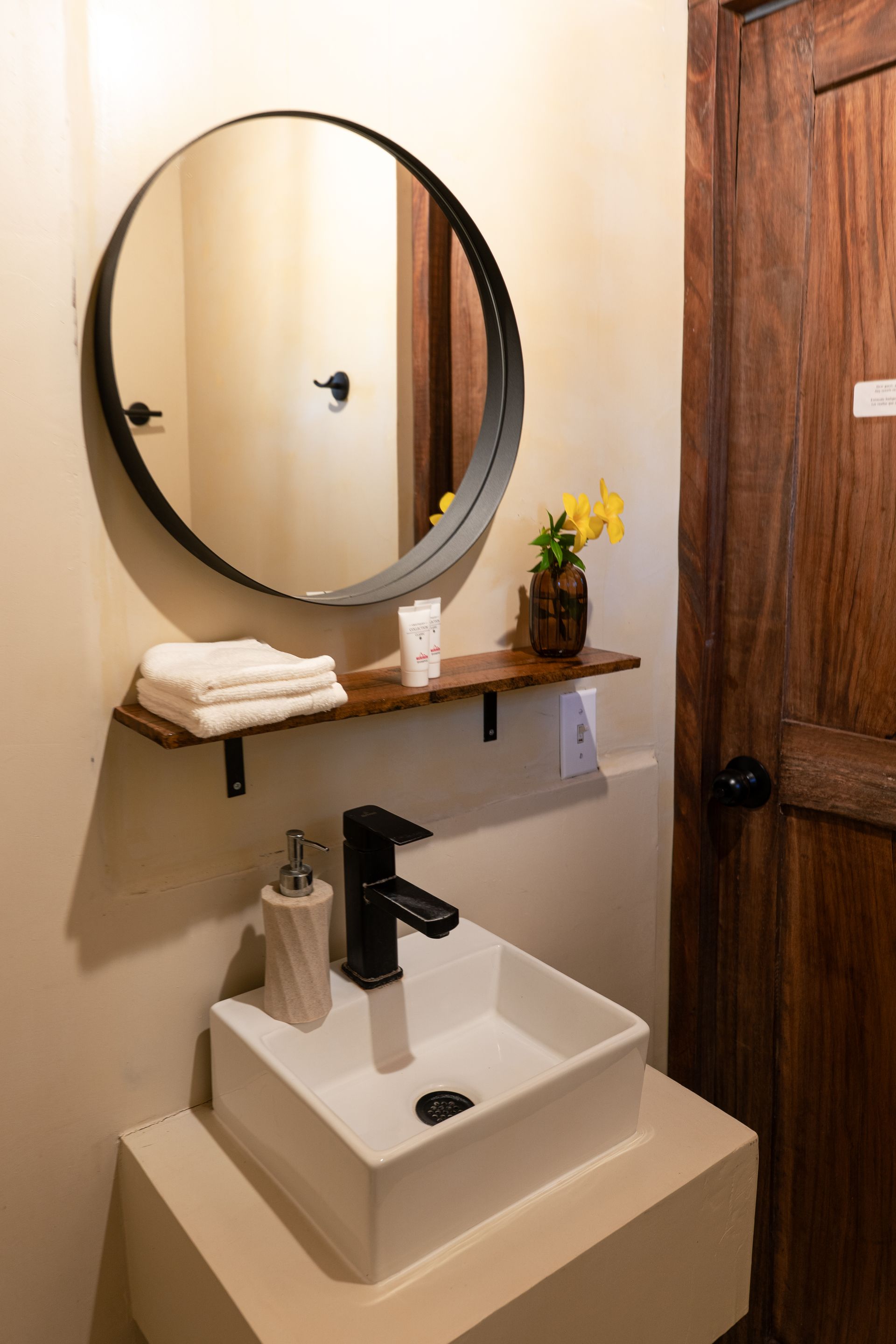 Small bathroom vanity with round mirror, white sink, black faucet, and wooden shelf with towels and flowers.