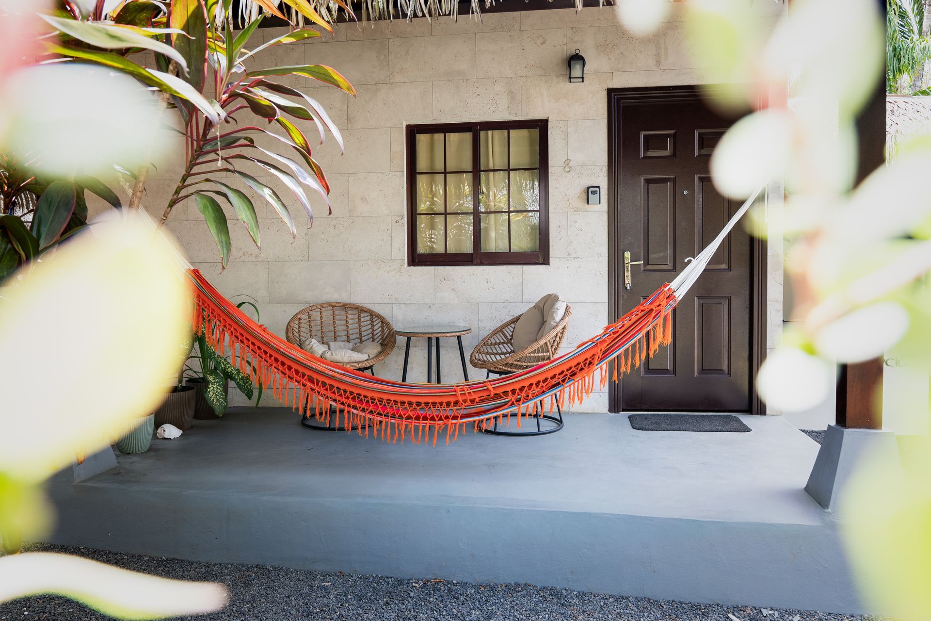 Front porch with orange hammock chairs, a small table, and a dark door beside a window with potted plants.
