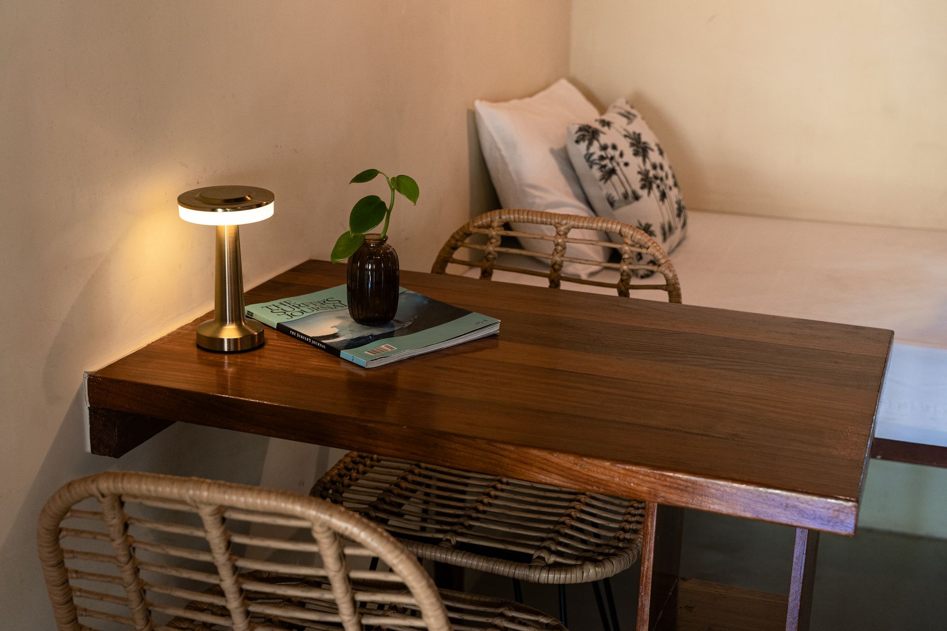 Cozy dining nook with wooden table, wicker chairs, small lamp, plant, and daybed by the wall